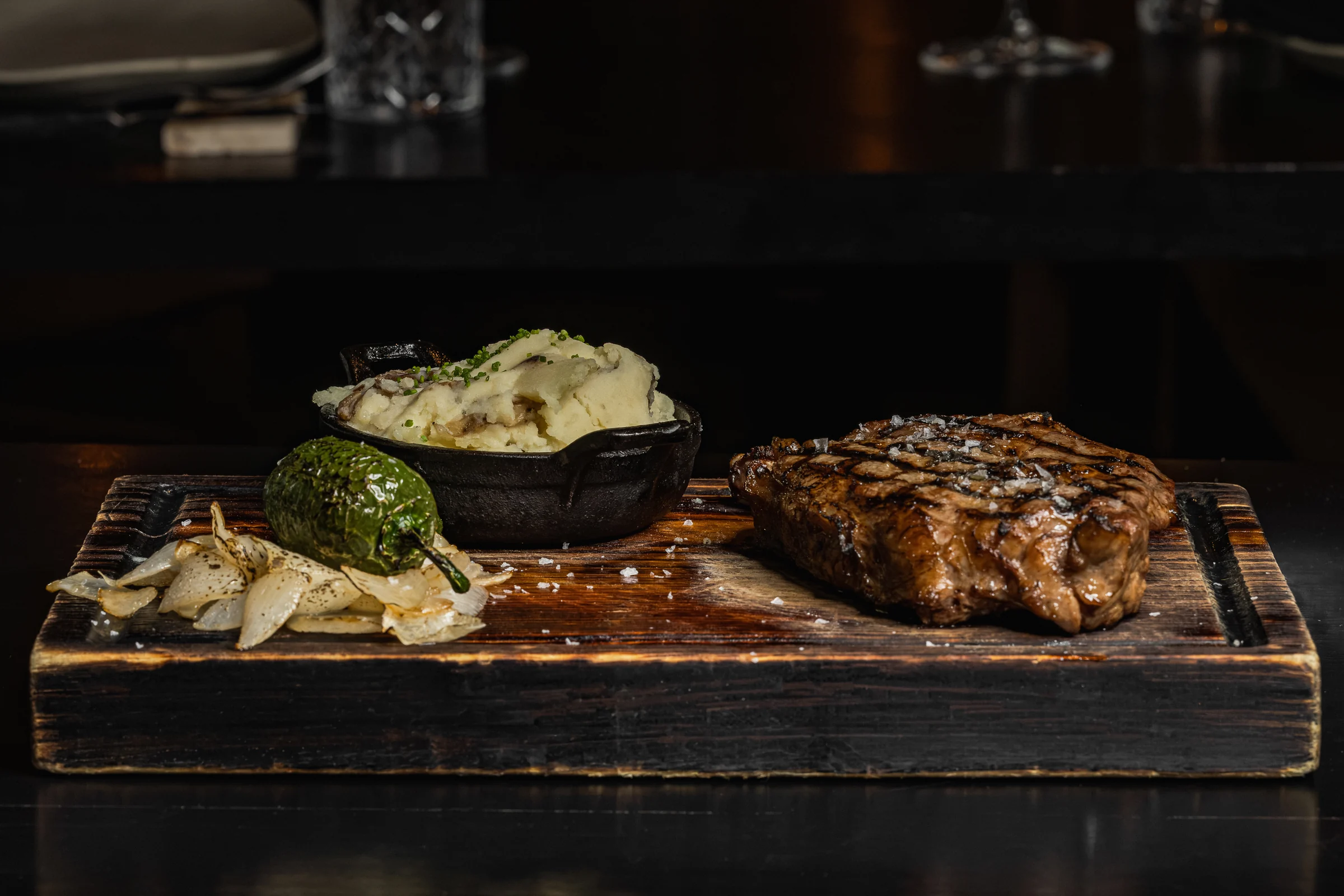 A mesquite-grilled steak on a charred wooden board with potatoes, roasted onions and a jalapeño at Origen