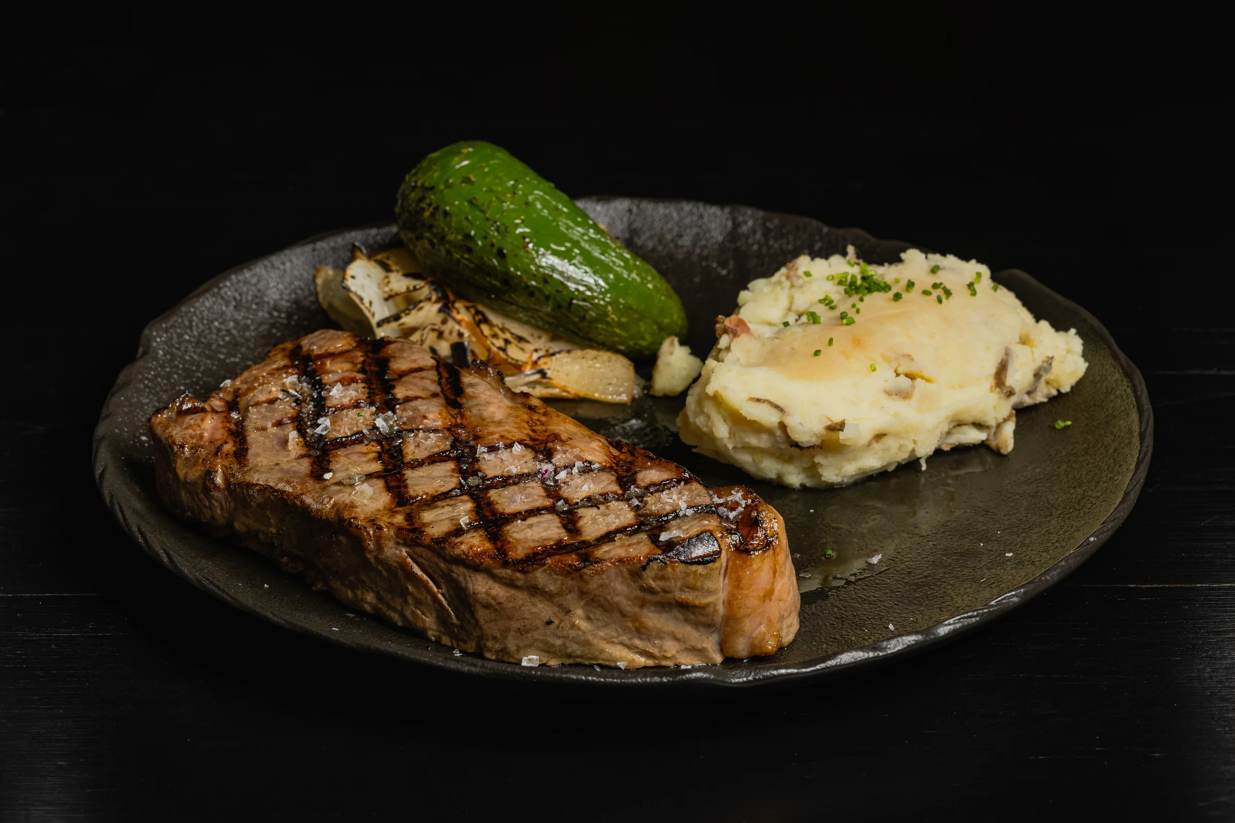 Grilled steak with mashed potatoes, onions and a roasted green pepper on a dark plate.